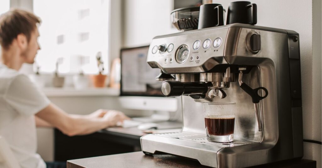 Close-up of a home Coffee machine pouring a coffee into a glass , with a person working at his desk behind it