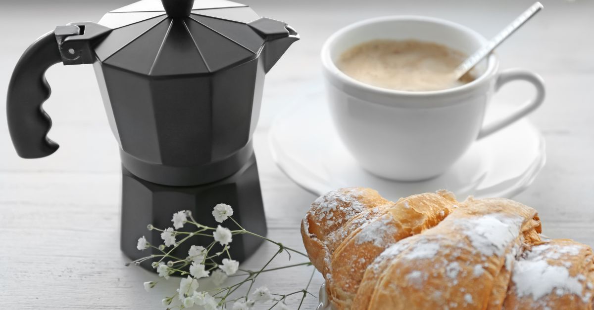 An Almond Croissant on the tablke with some Babies Breath Flowers. A cup of coffee and a moka pot