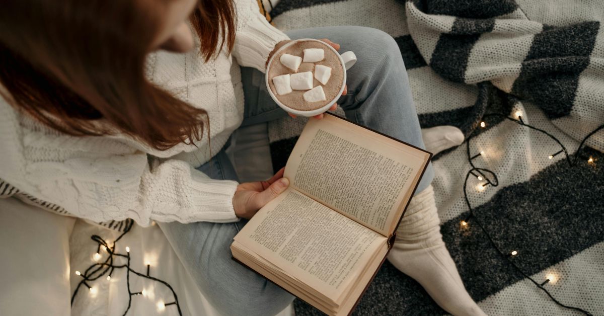 Person reading a book with a hot drink in her hand and some string lights around her